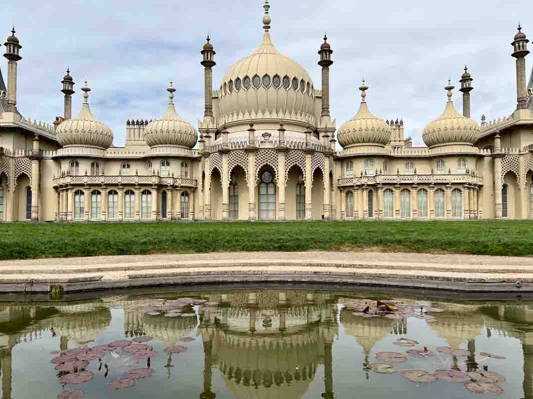 Brighton Royal Pavilion with a grey sky background. A cream onion domed building with pillars, arched windows, stone lattice work reflected ina pond with lily leaves. A visit to the Pavilion is one of the best things to do in Brighton. 