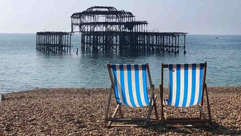 Brighton's West pier with two blue and white striped deckchairs looking towards it from the pebble beach