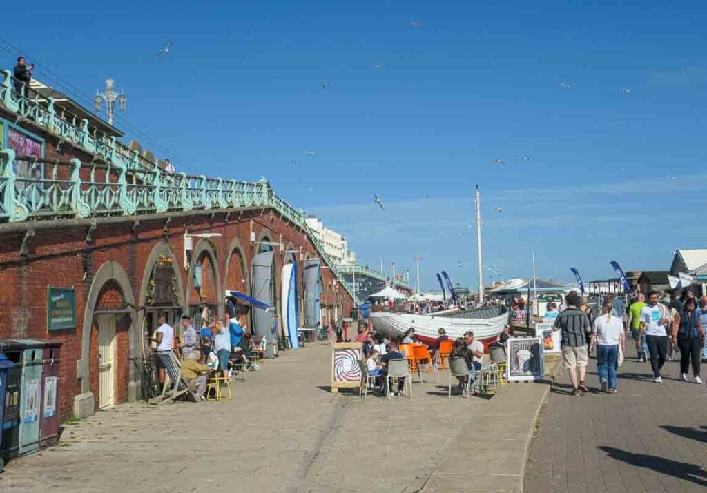 Brighton Seafront on a busy sunny day. People walk along the promenade and a fishing boat sits outside the Fishing Museum. On the left are the arches with people sitting on deckchairs. Seagulls fly in the air