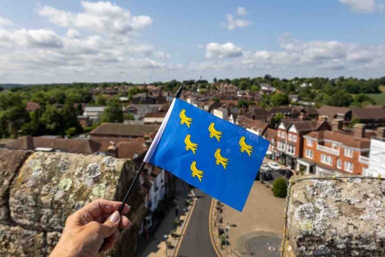 Waving a hand held Sussex flag which is a blue field with six golden martlets. In the background you can see a view of Battle High Street