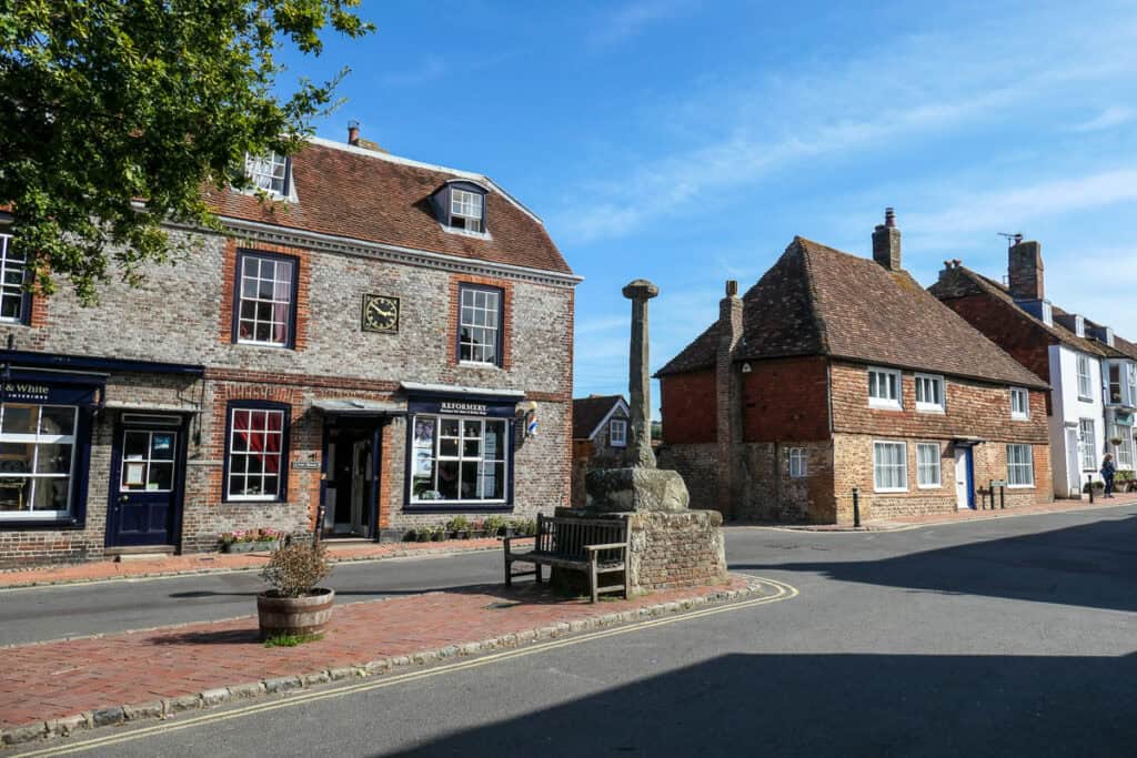 A stone memorial in Alfriston, East Sussex with flint house behind. It has a black door and six windows. There is a clock on the building which reads 2:50 pm. More old with buildings can be seen leading out of shot on the right of the image