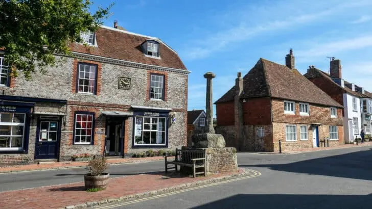 A stone memorial in Alfriston, East Sussex with flint house behind. It has a black door and six windows. There is a clock on the building which reads 2:50 pm. More old with buildings can be seen leading out of shot on the right of the image