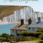 Coastguard cottages at Seaford Head with the Seven Sisters white cliffs and a calm blue English channel in the background. A path leads down to the cottages which are partly obscured by shrubs. Text overlay reads the best things to do in Sussex, England