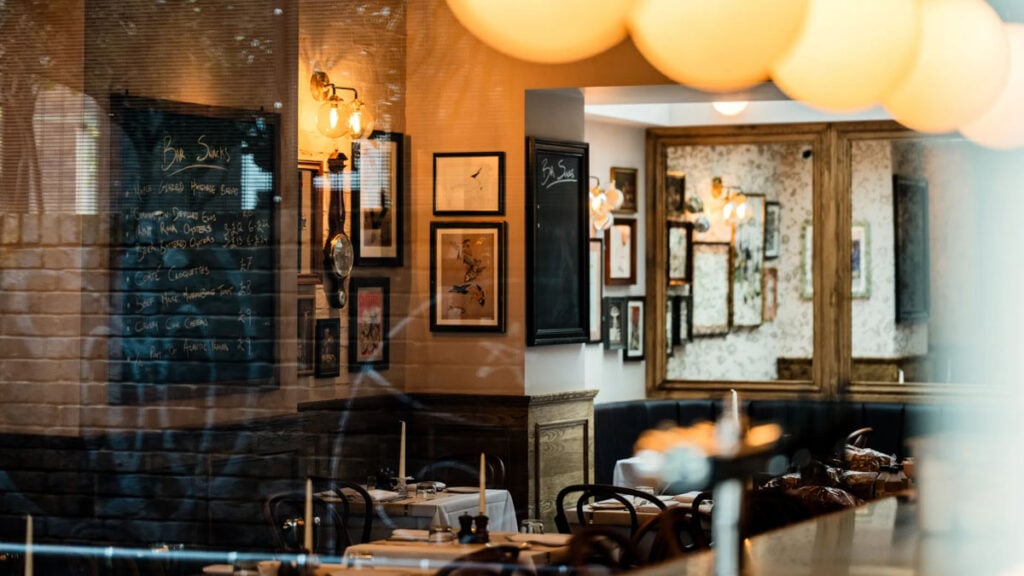 Crazy Goose, Brighton. Looking through a window into a warm cozy bar and restaurant with candles on tables and pictures on the walls. A blackboard on the wall has the bar snacks chalked on it