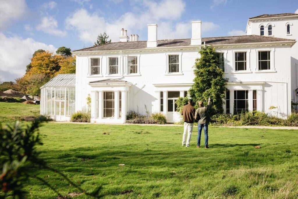 Long Lane Hotel, West Sussex. A white country house hotel with two men standing looking at it from the lawn in front. Blue sky and fluffy clouds are behind
