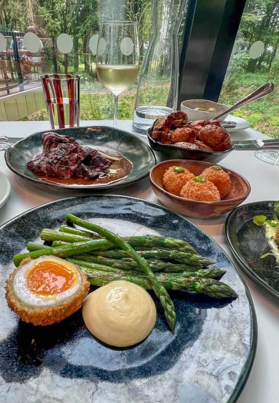 Small plates on a table with a glass of white wine. Plates include half a scotch egg with mayonnaise and asparagus, 3 arancini, roasted new potatoes and beef bavette
