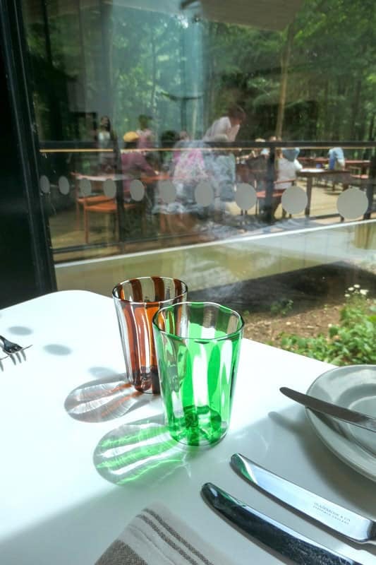 24 the restaurant at Goodwood Art Foundation. Two striped water glasses on a table with sunlight reflecting through the glass onto the table. A terrace with people dining is in the background