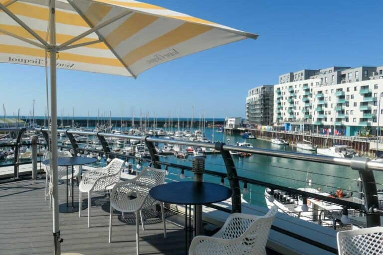 Terrace overlooking Brighton Marina with boats in the water and a building in the background. A yellow and white striped parasol is on the terrace along with chairs and tables
