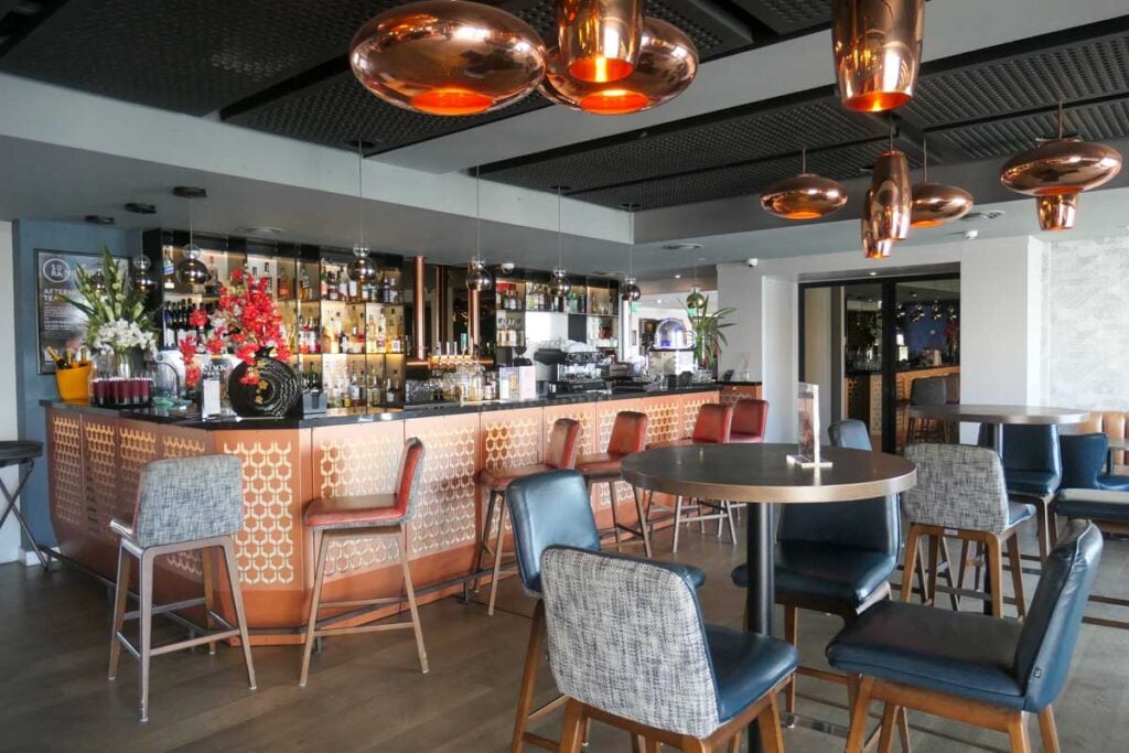 The bar in Malmaison Brighton Marina. Copper lights hang from the ceiling, five bar stools are next to the bar and there are tables and chairs in the room. The bar has bottles and glasses at the back with a display of red flowers on it