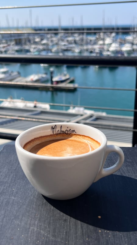 A white china cup with a flat white coffee. The word Malmaison is printed on the inside of the cup. Brighton marina is in the background