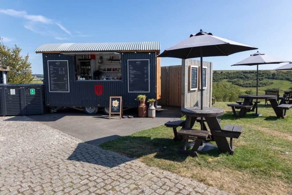 A shepherd's hut is used as a food stall next to picnic tables with parasols in a countryside setting with blue sky