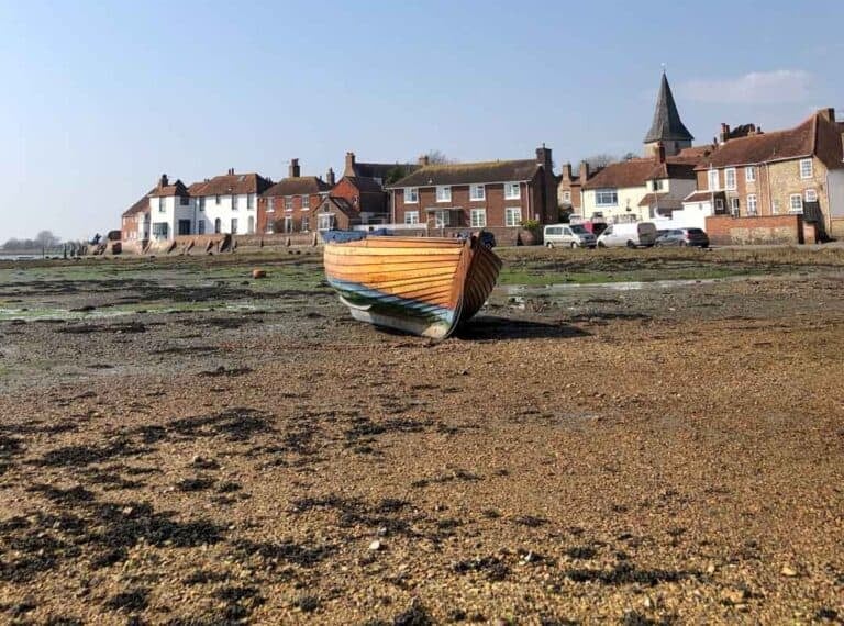 A boat sits on the single at Bosham in West Sussex. In the back ground are houses lining the estuary with the church spire in the background