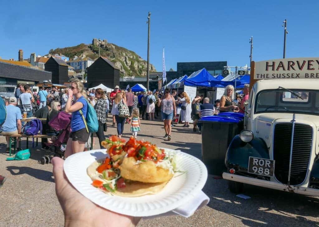 A hand holds out a paper plate with seafood and salad. A food festival is happening in the background.